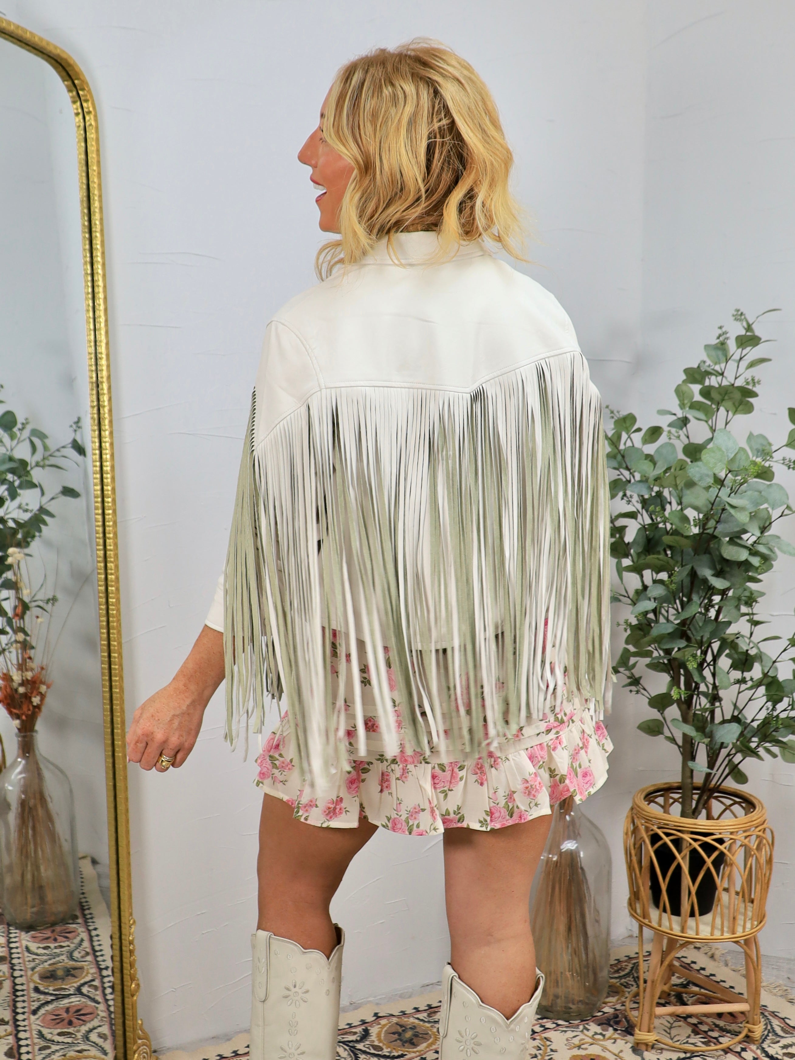 Woman wearing white faux leather fringe jacket with floral hem detail, viewed from behind, styled with white cowboy boots.
