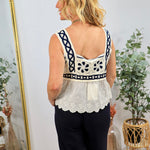 Woman wearing white embroidered sleeveless top with navy blue trim and scalloped hem, paired with black pants, shown from beh