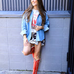 Woman wearing oversized light wash denim shirt layered over graphic tee and cow print mini skirt with red cowboy boots