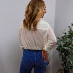 Woman wearing a sheer cream long-sleeve blouse tucked into blue wide-leg jeans, back view.