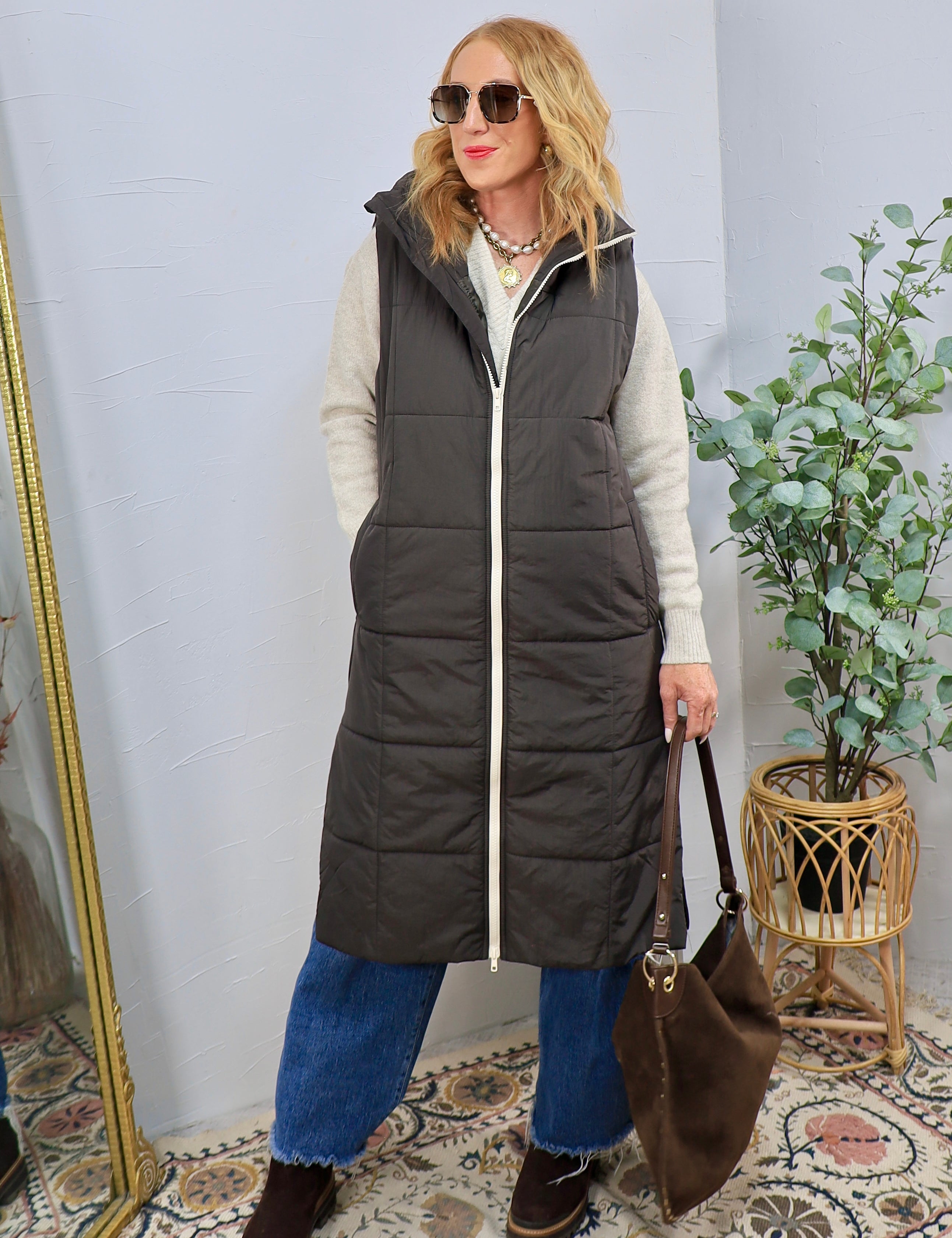 Woman wearing a long charcoal quilted puffer vest over a cream sweater with jeans and brown boots