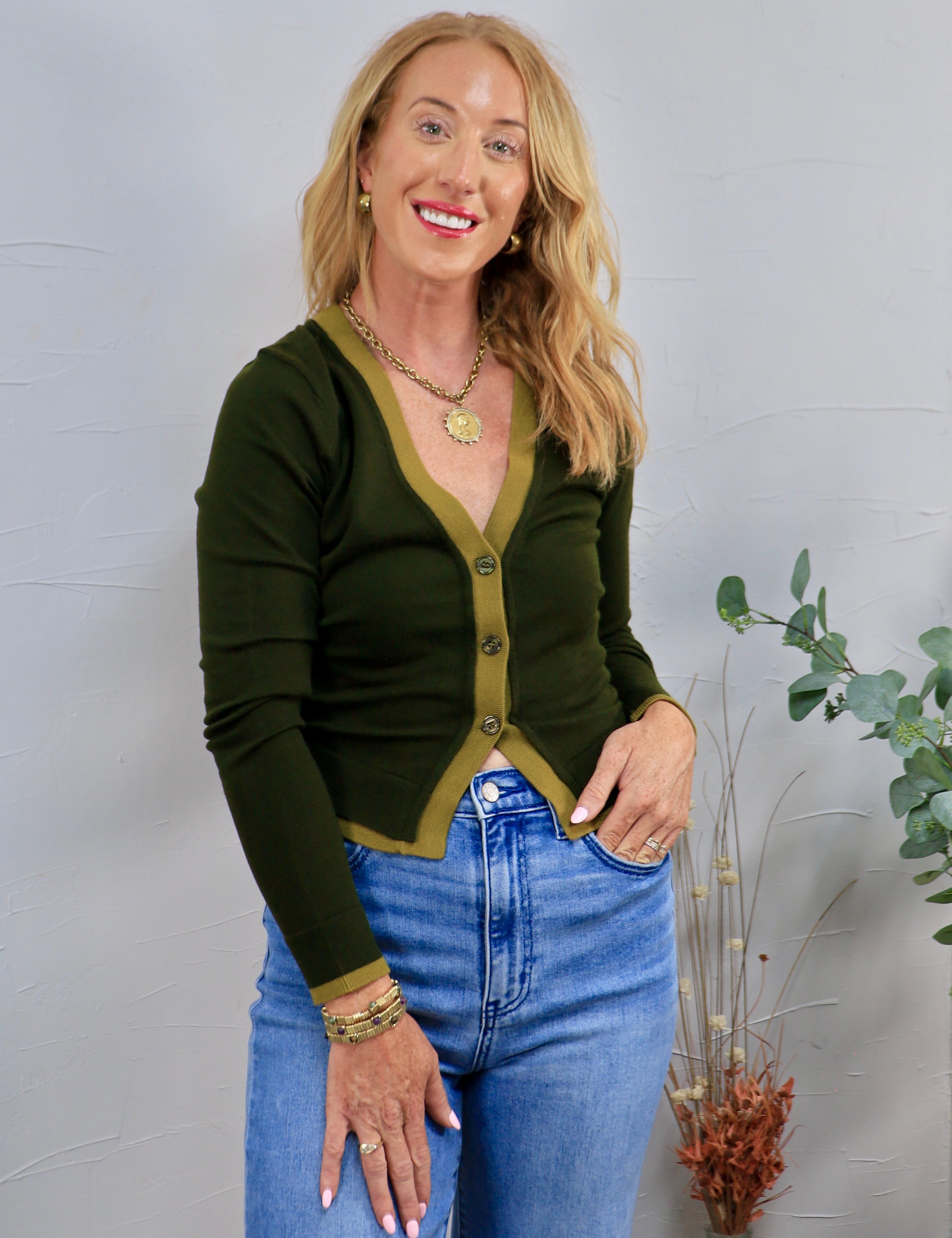 Dark olive green cardigan with contrast trim, gold buttons, styled with blue high-waist jeans and gold jewelry.