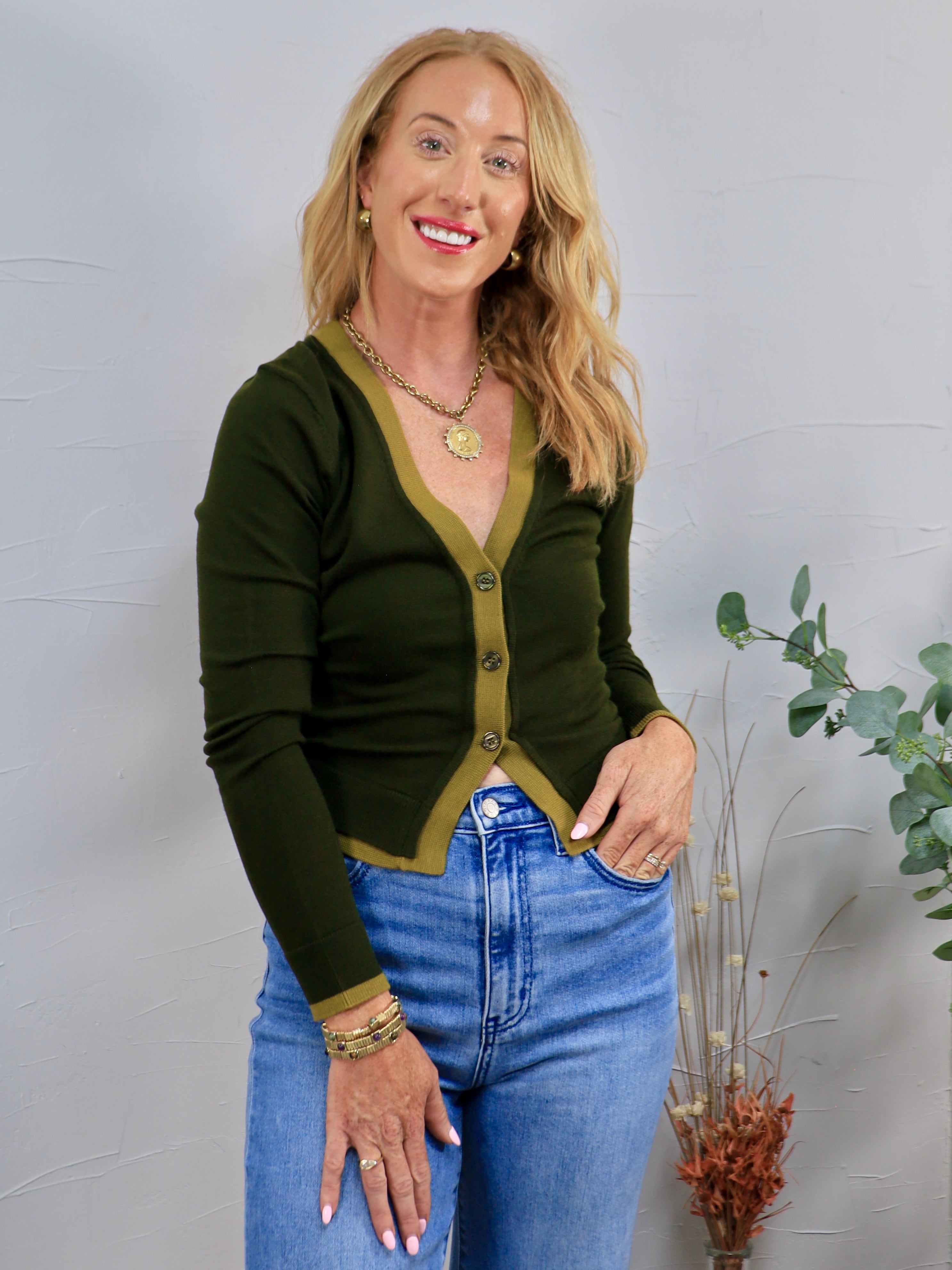 Dark olive green cardigan with contrast trim, gold buttons, styled with blue high-waist jeans and gold jewelry.
