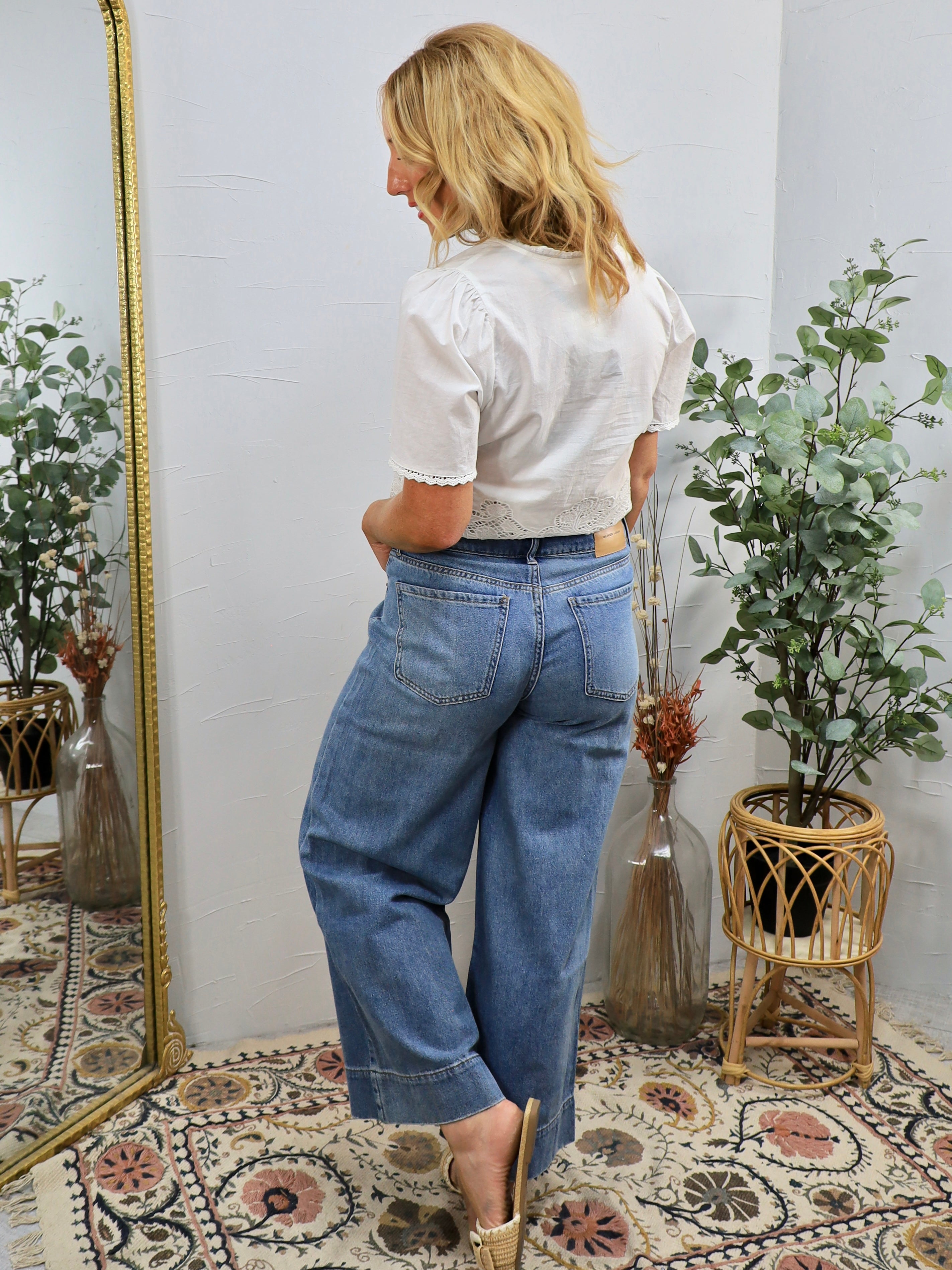 Woman wearing light blue vintage-style wide-leg cropped jeans with white lace-trimmed blouse, viewed from behind in boho-styl