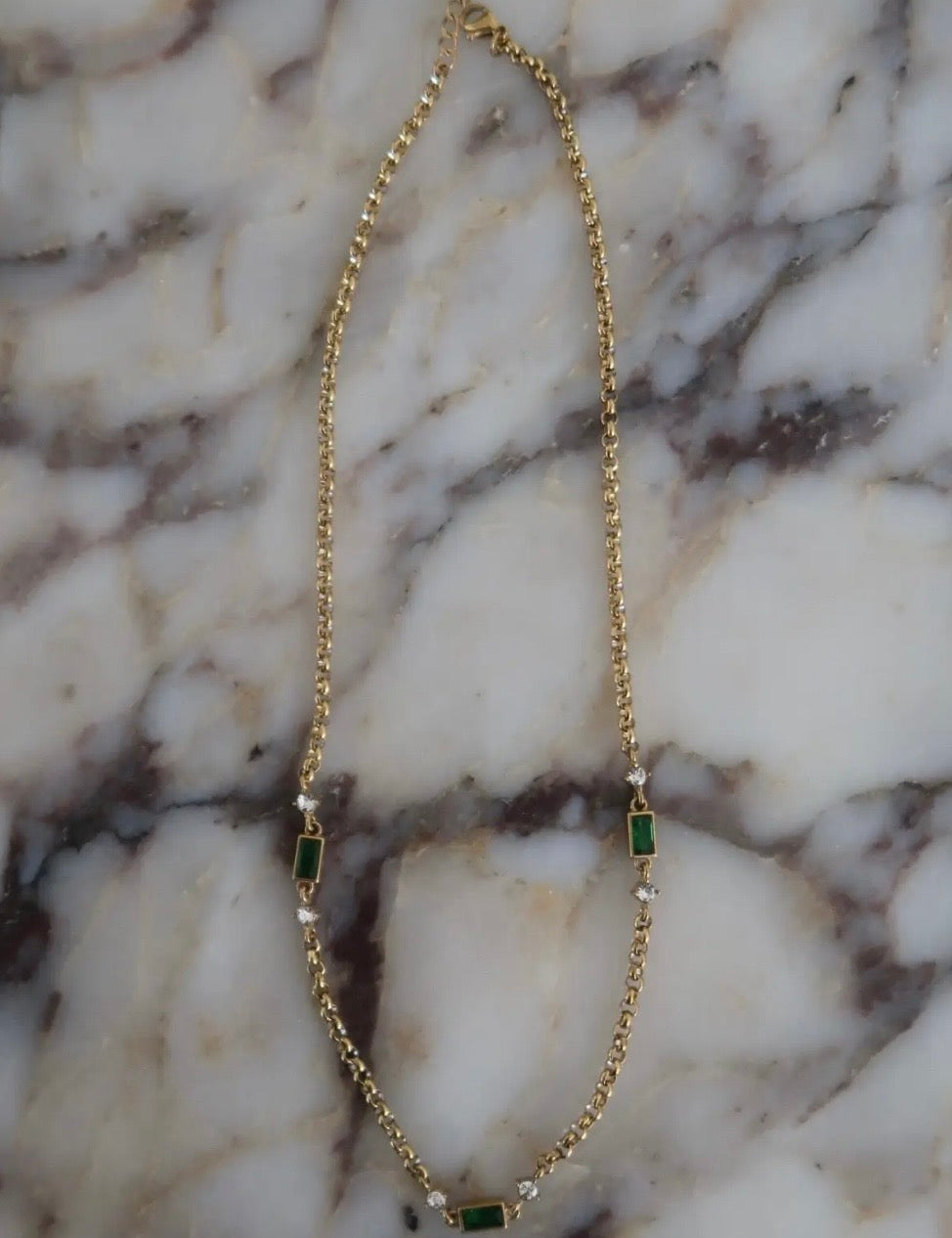 Gold rolo chain necklace with emerald green baguette stones and clear crystal accents on marble surface