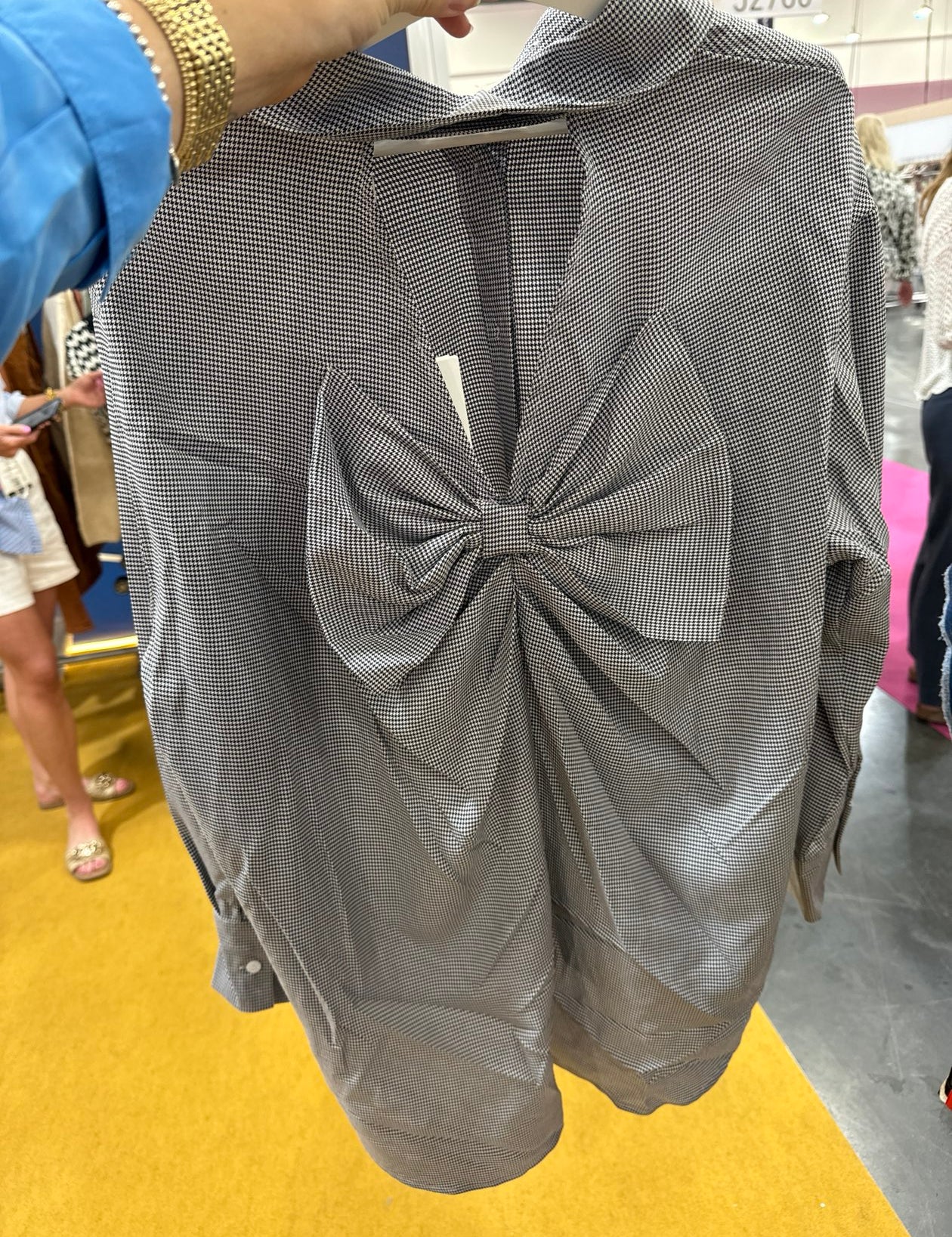 Gray houndstooth patterned blouse with large bow detail at front, long sleeves, being held up in store setting.