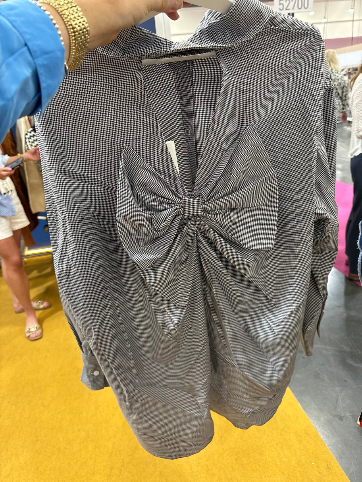 Gray houndstooth patterned blouse with large bow detail at front, long sleeves, being held up in store setting.