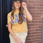 Woman wearing a camo trucker hat with gold Champs lettering, smiling against a brick wall