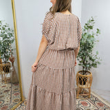 Woman wearing brown and white gingham checkered tiered midi dress with short sleeves, cinched waist, and ruffled layers.