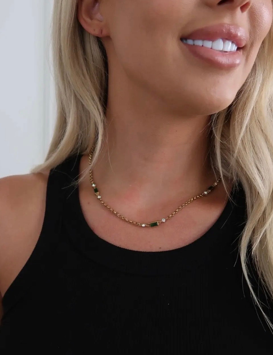Gold chain necklace with green baguette stones and crystal accents, worn on a model in a black tank top.