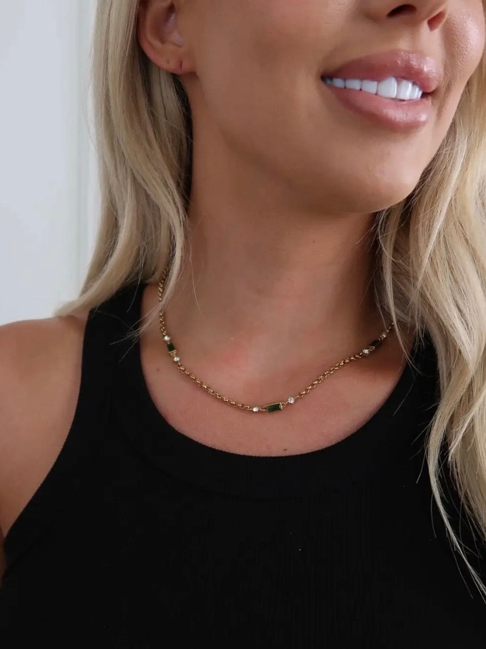 Gold chain necklace with green baguette stones and crystal accents, worn on a model in a black tank top.