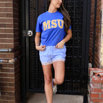 Blue MSU graphic tee with gold lettering, styled with blue gingham lace-trim shorts and white sneakers.