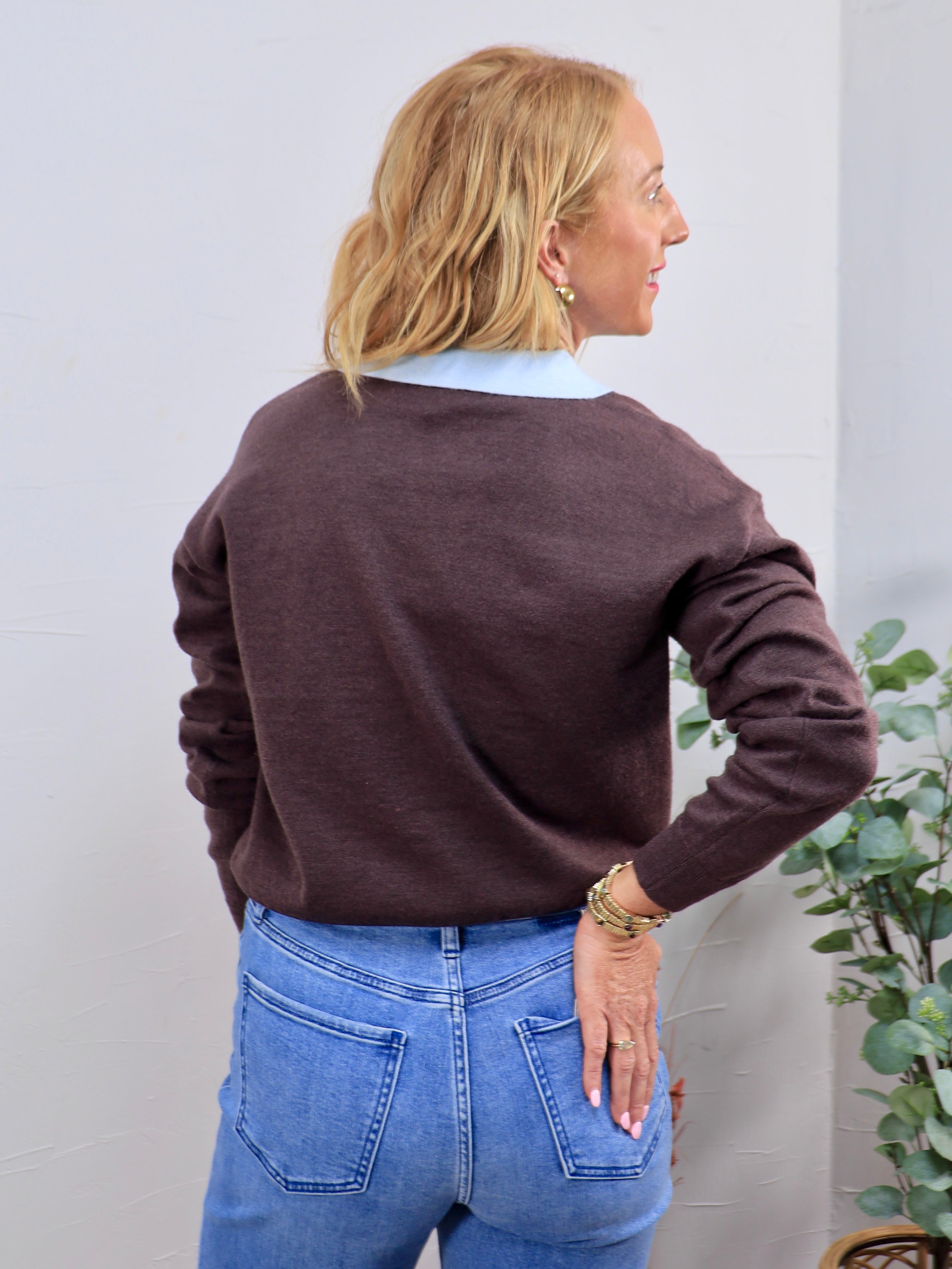 Brown knit sweater layered over light blue collared shirt, paired with blue jeans, back view.