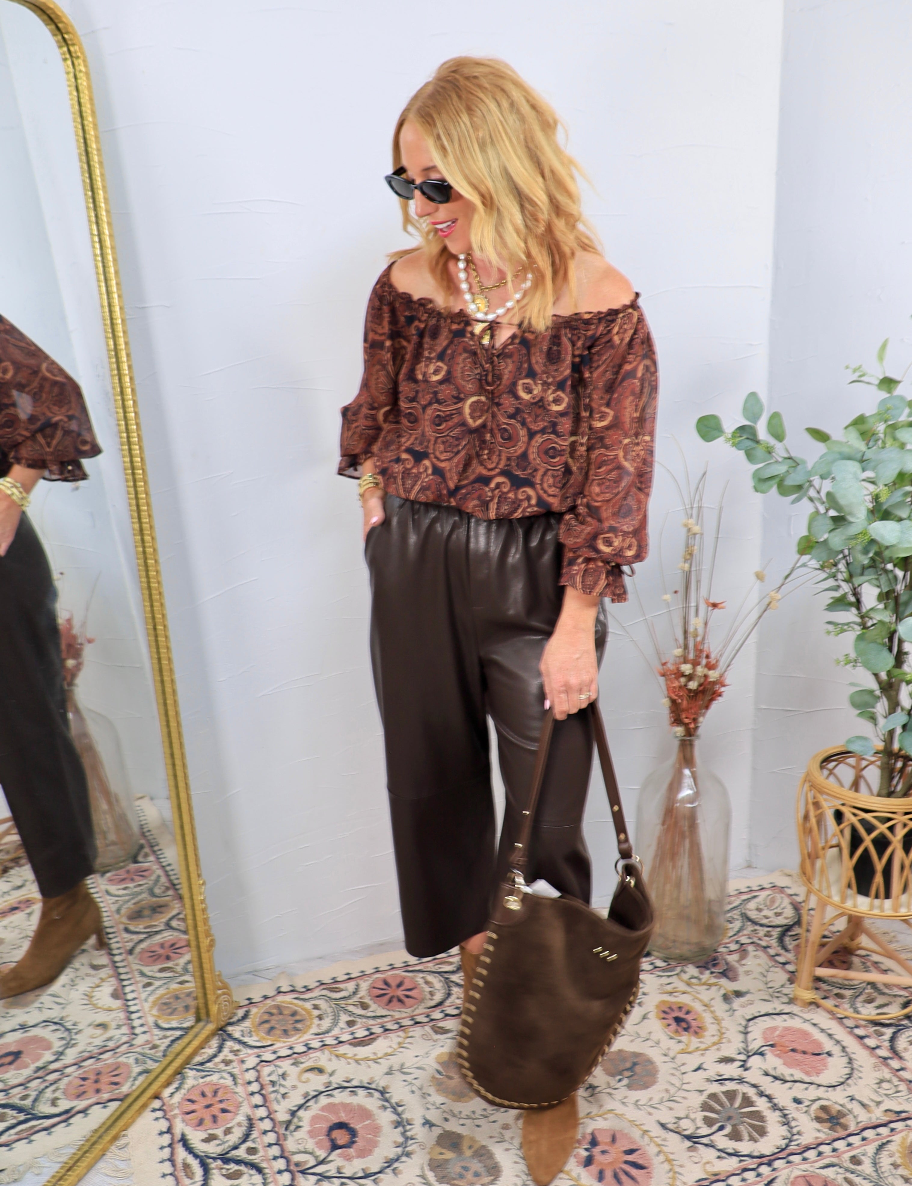 Dark brown faux leather wide-leg pants styled with paisley off-shoulder top, brown boots, and matching tote bag.