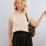 Woman wearing a beige sleeveless sweater and black skirt, holding a brown handbag with gold studs against a light gray background.