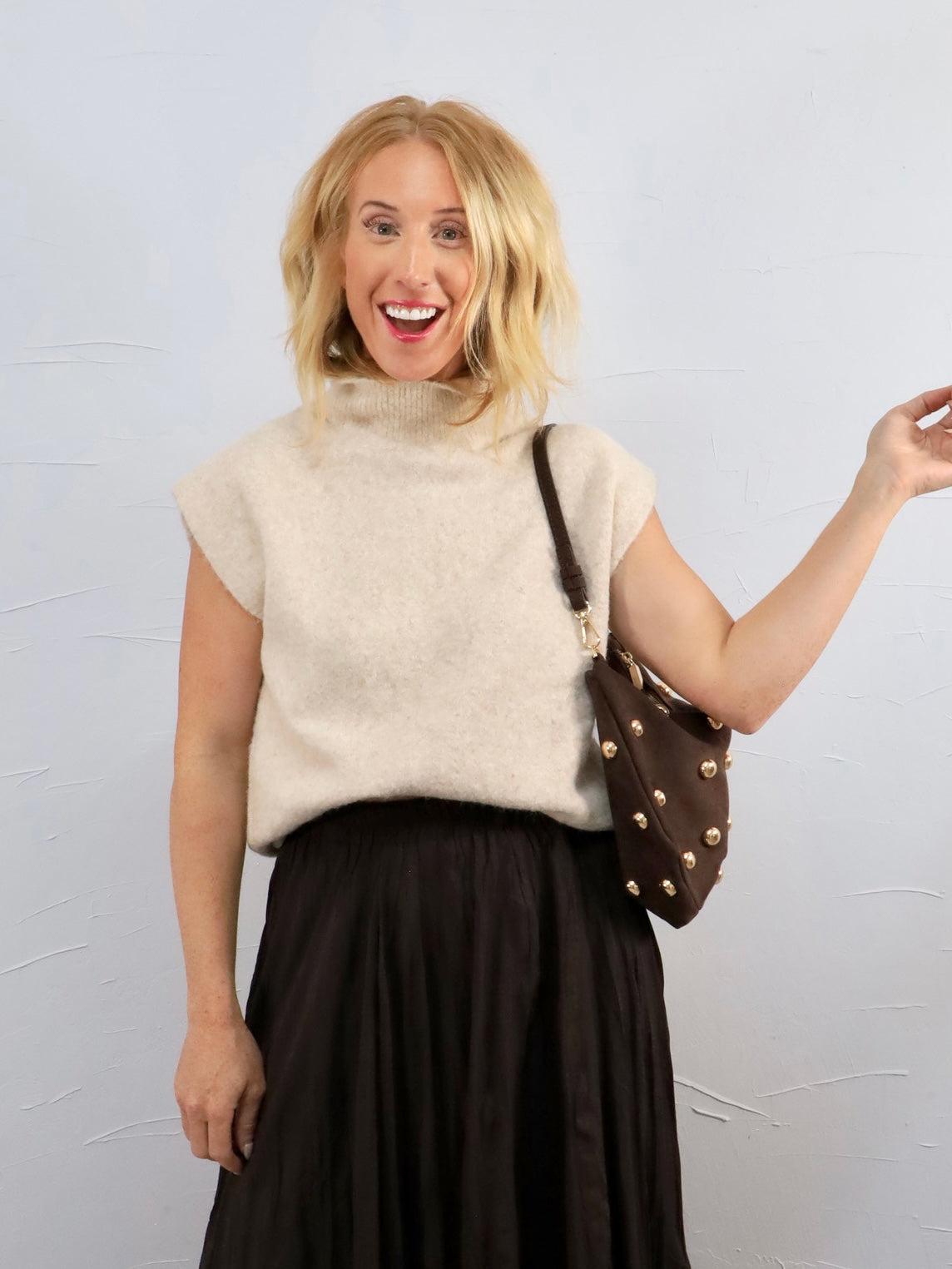 Woman wearing a beige sleeveless sweater and black skirt, holding a brown handbag with gold studs against a light gray background.