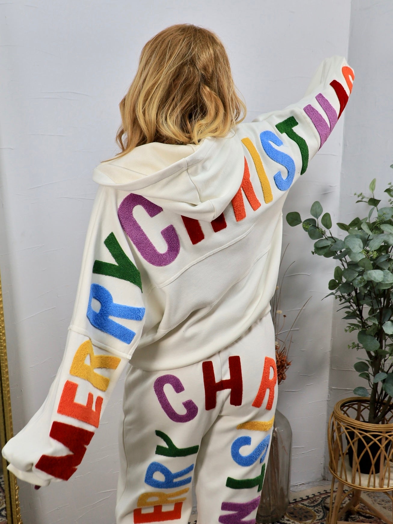 White hoodie and jogger set with colorful "Merry Christmas" lettering, modeled from behind, arms outstretched.