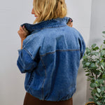 Woman modeling back view of oversized medium-wash blue denim bomber jacket with elastic hem, paired with brown mini skirt.