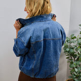 Woman modeling back view of oversized medium-wash blue denim bomber jacket with elastic hem, paired with brown mini skirt.