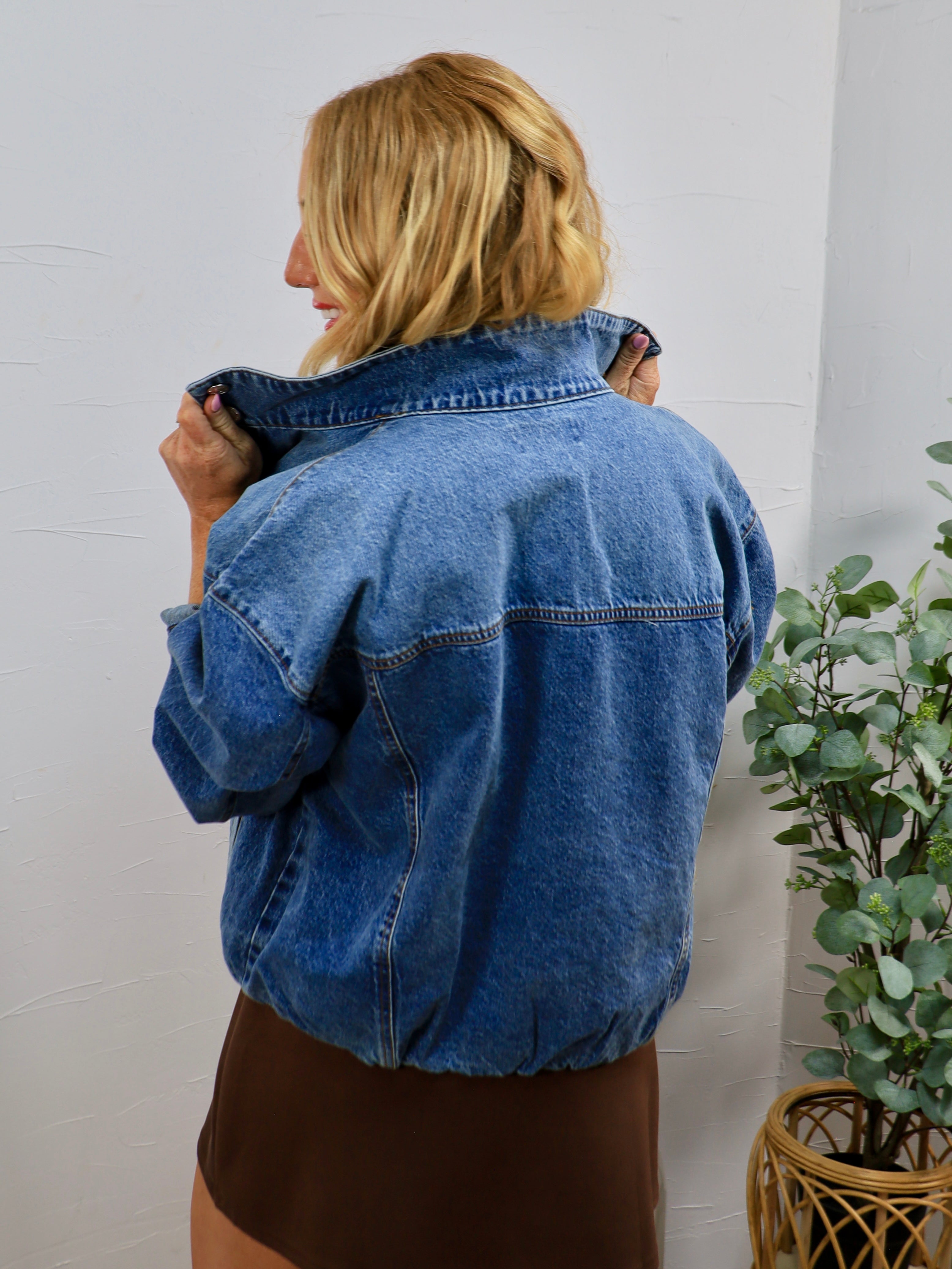 Woman modeling back view of oversized medium-wash blue denim bomber jacket with elastic hem, paired with brown mini skirt.