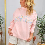 Woman wearing pink mockneck sweatshirt with colorful floral patchwork "MONTANA" text, styled with white shorts in boho interi