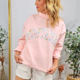 Woman wearing pink mockneck sweatshirt with colorful floral patchwork "MONTANA" text, styled with white shorts in boho interi