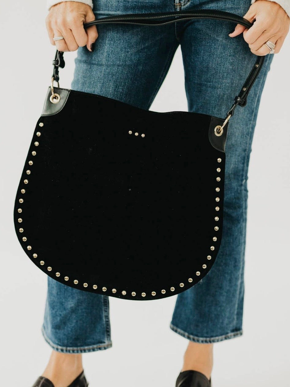Black suede hobo tote bag with gold stud trim and adjustable strap, held by model wearing blue jeans.