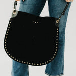 Black suede hobo tote bag with gold stud trim and adjustable strap, held by model wearing blue jeans.