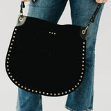 Black suede hobo tote bag with gold stud trim and adjustable strap, held by model wearing blue jeans.