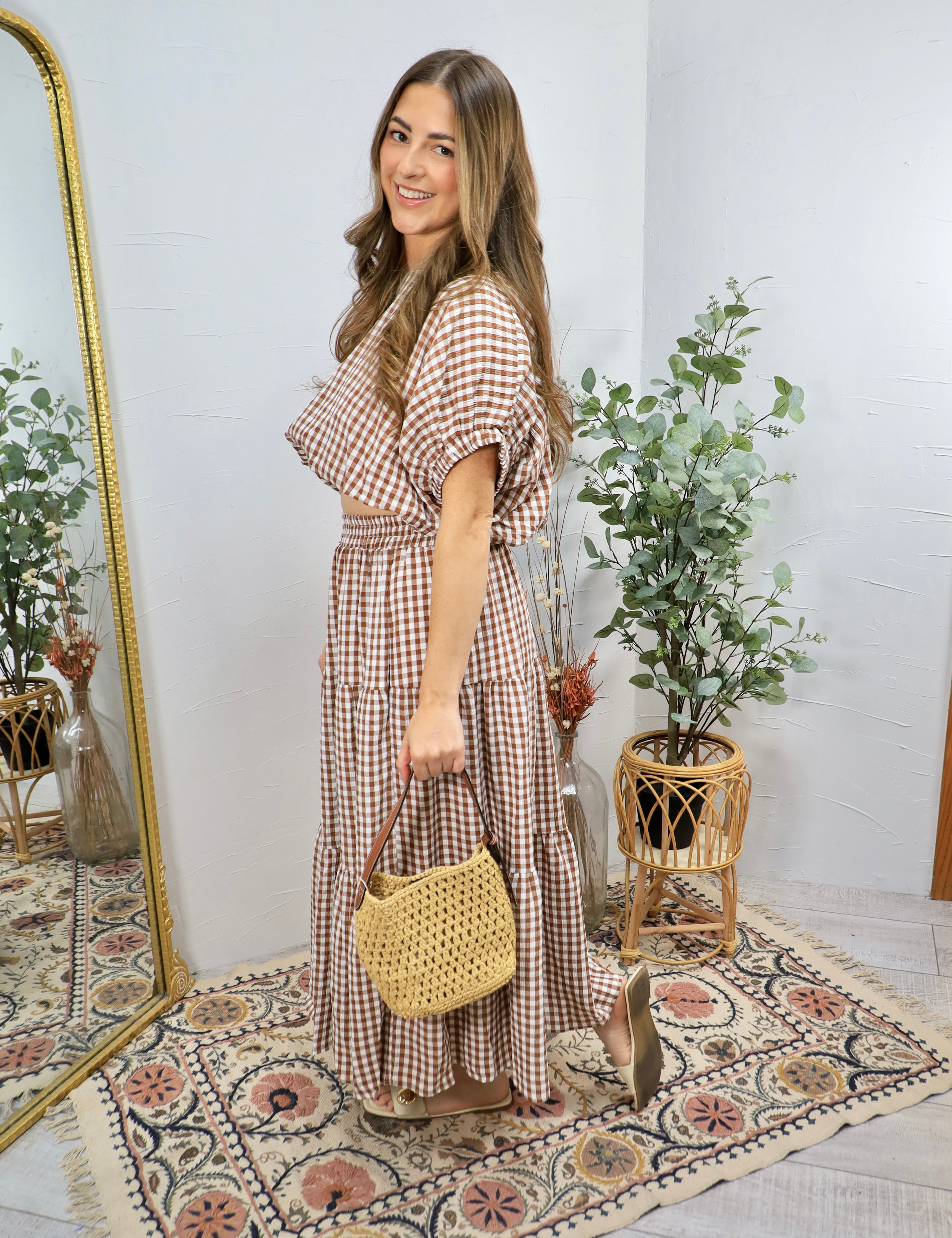Woman wearing brown and white gingham checkered tiered midi dress with puffed sleeves, holding woven straw handbag and nude h
