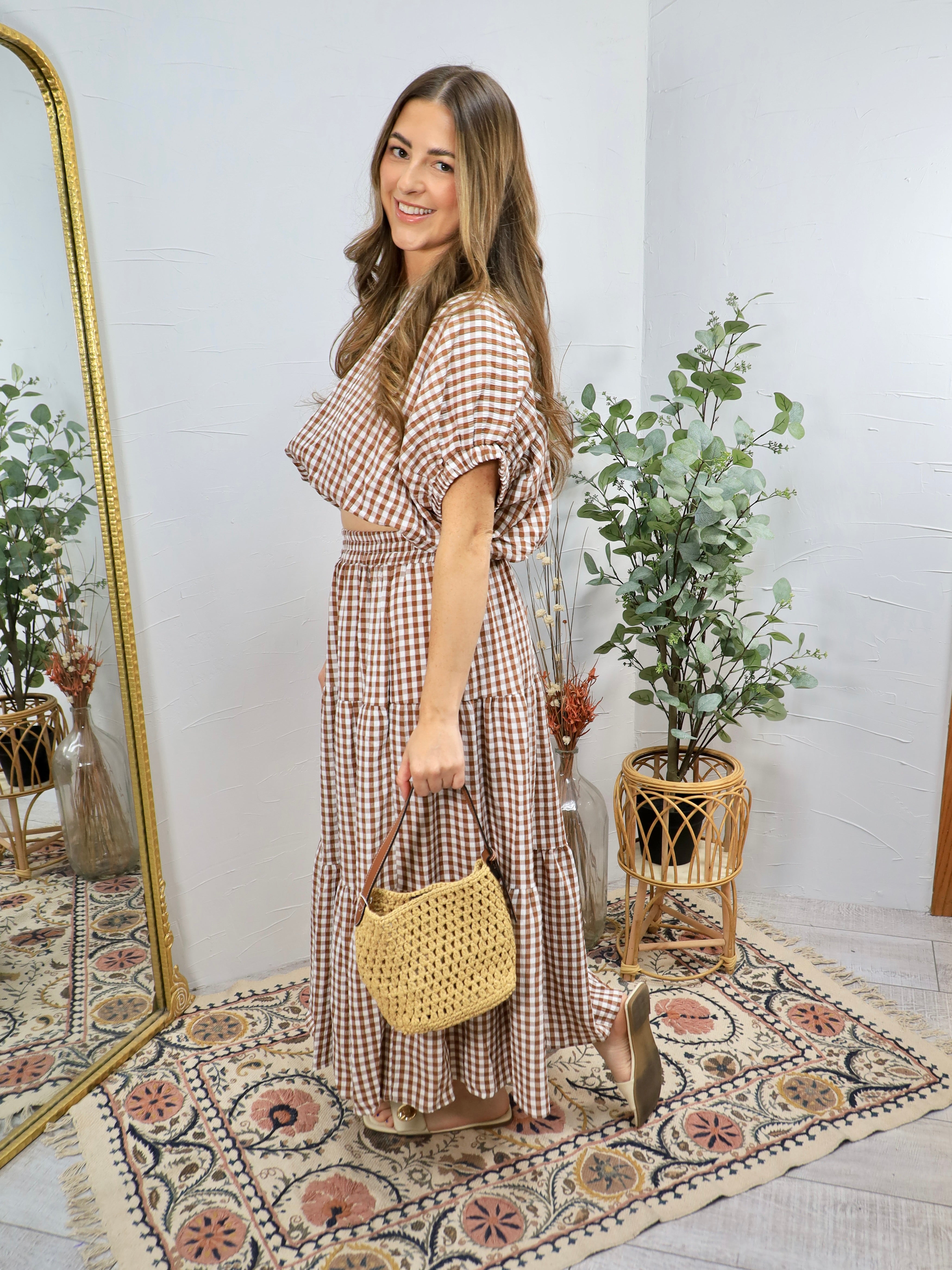 Woman wearing brown and white gingham checkered tiered midi dress with puffed sleeves, holding woven straw handbag and nude h