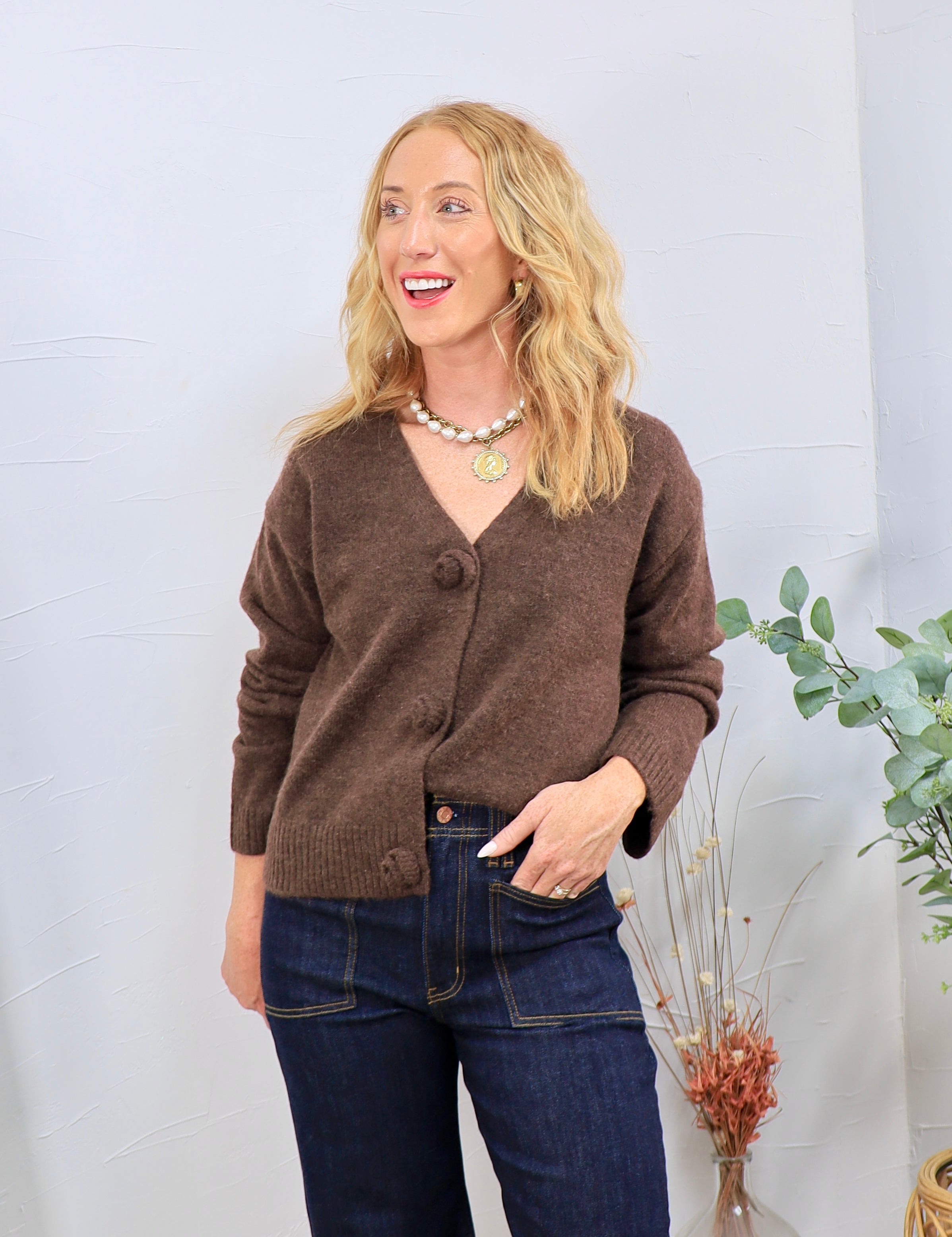 Brown fuzzy knit cardigan with pom-pom buttons, V-neckline, styled with dark wash straight-leg jeans.