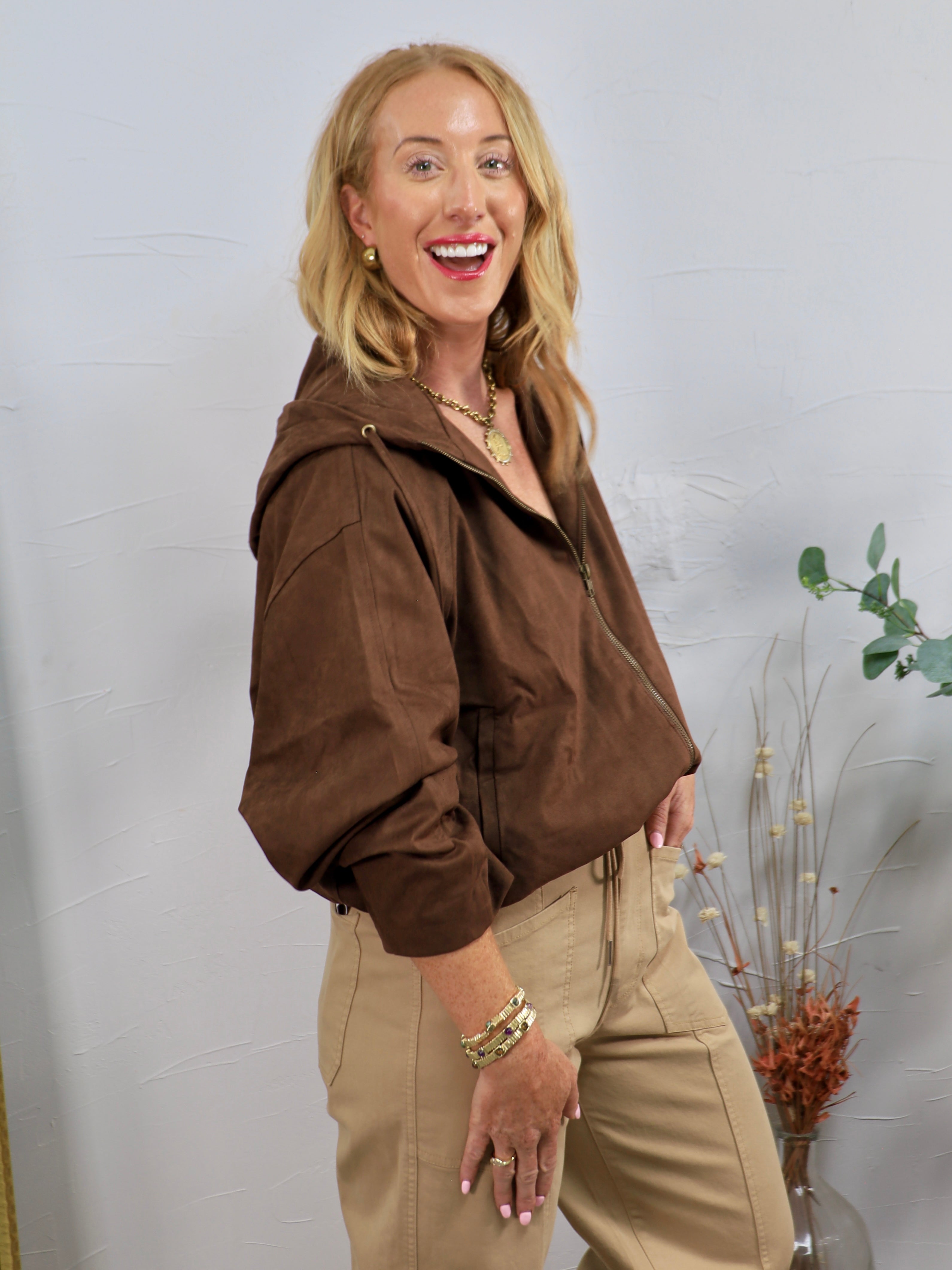 Chocolate brown hooded bomber jacket with zip front, styled with khaki trousers and gold jewelry.