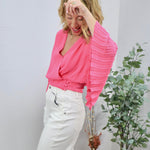 Pink wrap blouse with pleated sleeves, styled with white wide-leg jeans, modeled by blonde woman laughing.