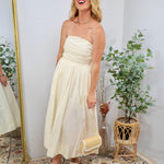 Woman wearing a strapless butter cream midi dress with ruched bodice, holding a woven clutch