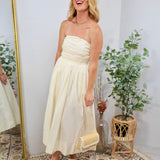Woman wearing a strapless butter cream midi dress with ruched bodice, holding a woven clutch