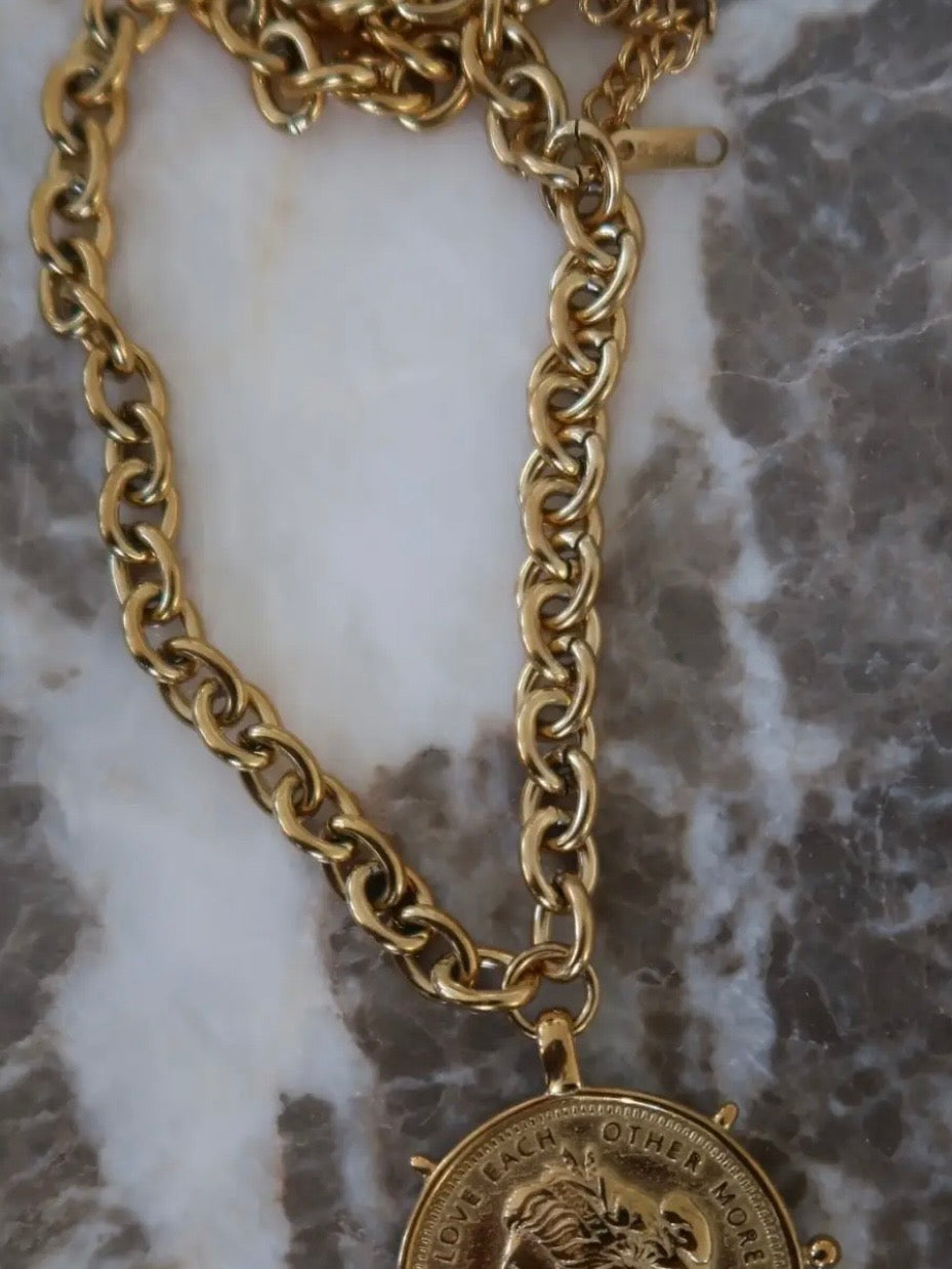 Gold chunky chain necklace with circular coin pendant engraved "Love Each Other More" on marble surface.
