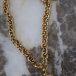 Gold chunky chain necklace with circular coin pendant engraved "Love Each Other More" on marble surface.