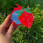 Strawberry-shaped compact mirror in red acrylic with green leaf detail and circular blue mirror insert.