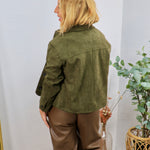 Olive green suede jacket worn from behind, styled with brown leather wide-leg trousers, button cuffs visible.