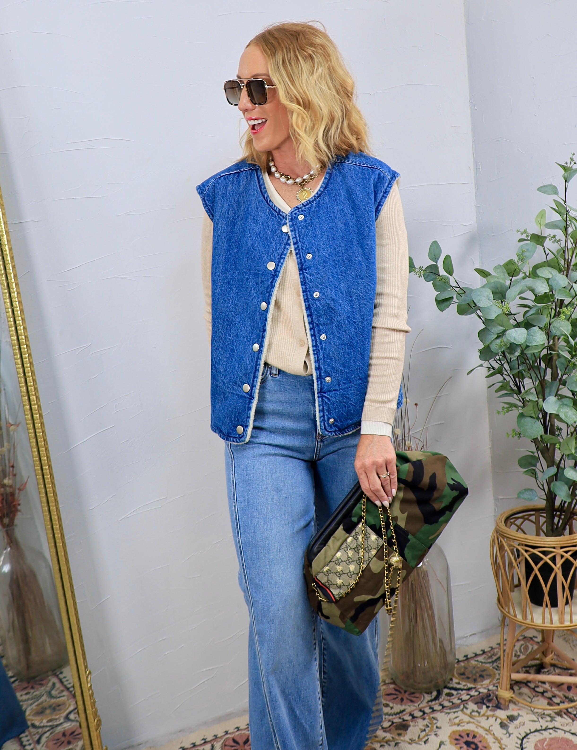 Woman wearing a blue denim sherpa-lined snap-front vest over a cream long-sleeve top with wide-leg jeans