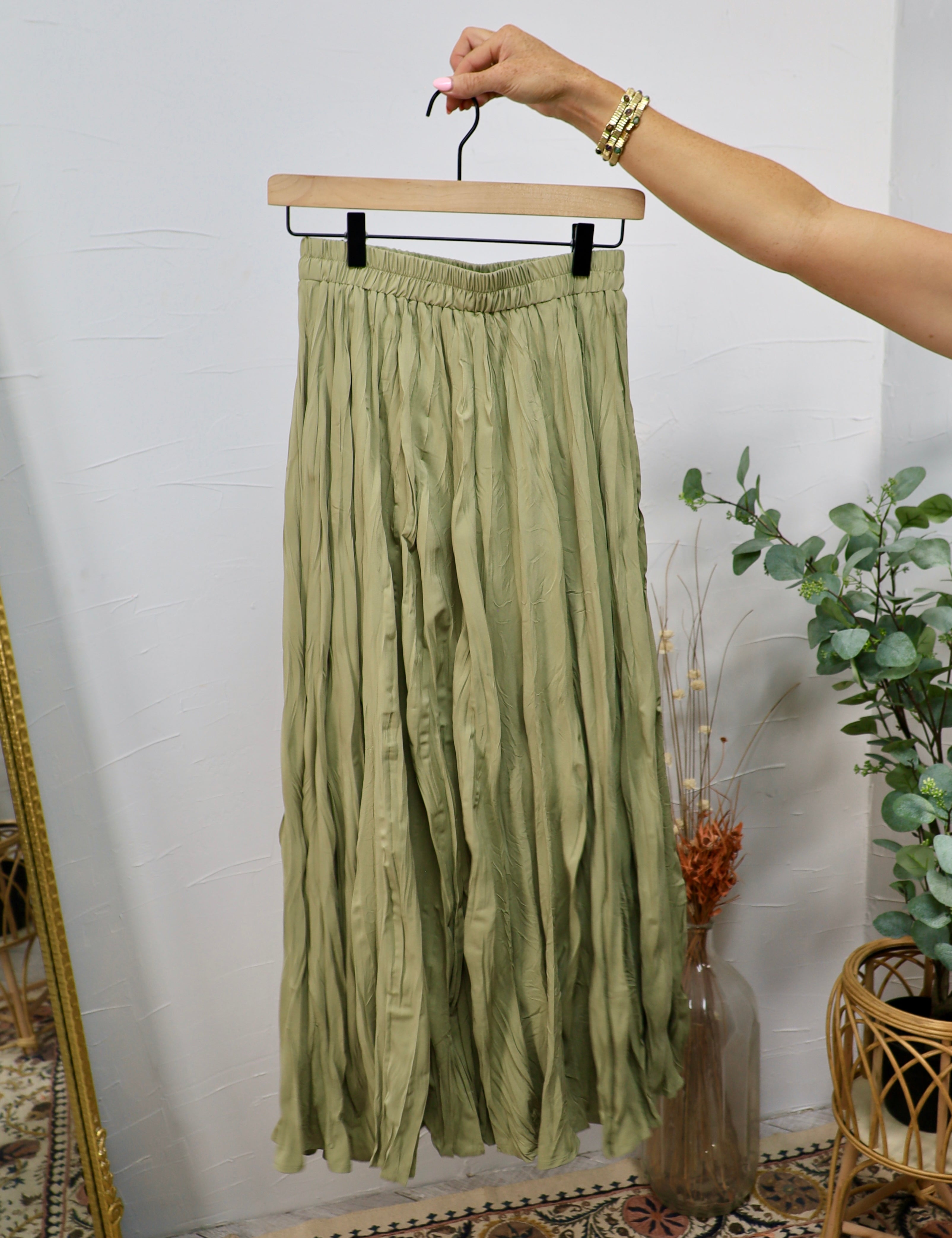 Olive green crinkled midi skirt with elastic waistband displayed on a wooden hanger against a white wall.