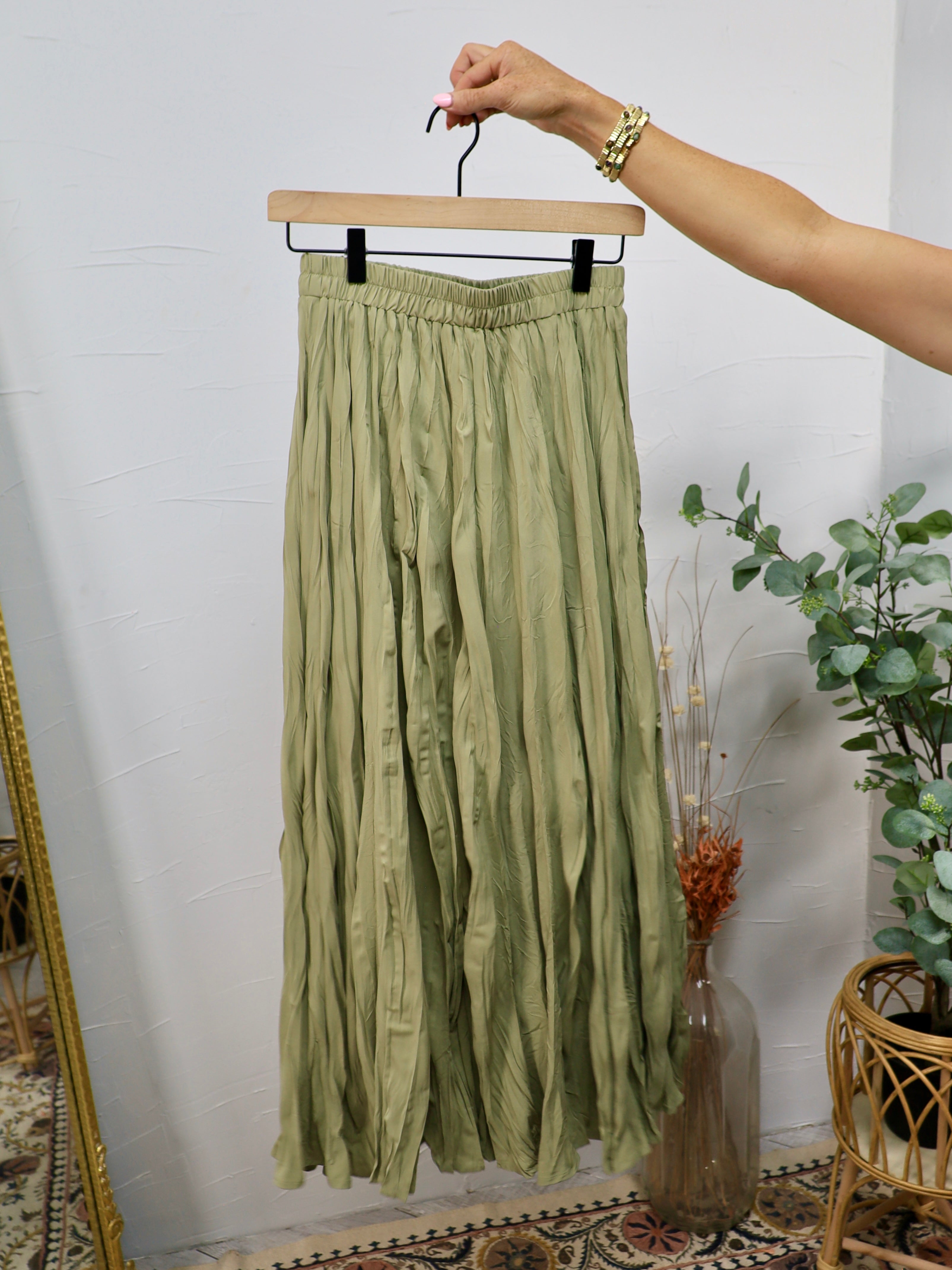 Olive green crinkled midi skirt with elastic waistband displayed on a wooden hanger against a white wall.