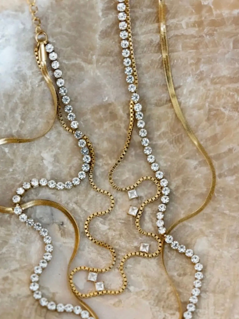 Gold tennis necklace with crystal rhinestones, box chain, and herringbone chain layered on marble surface.