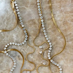 Gold tennis necklace with crystal rhinestones, box chain, and herringbone chain layered on marble surface.