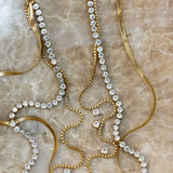 Gold tennis necklace with crystal rhinestones, box chain, and herringbone chain layered on marble surface.