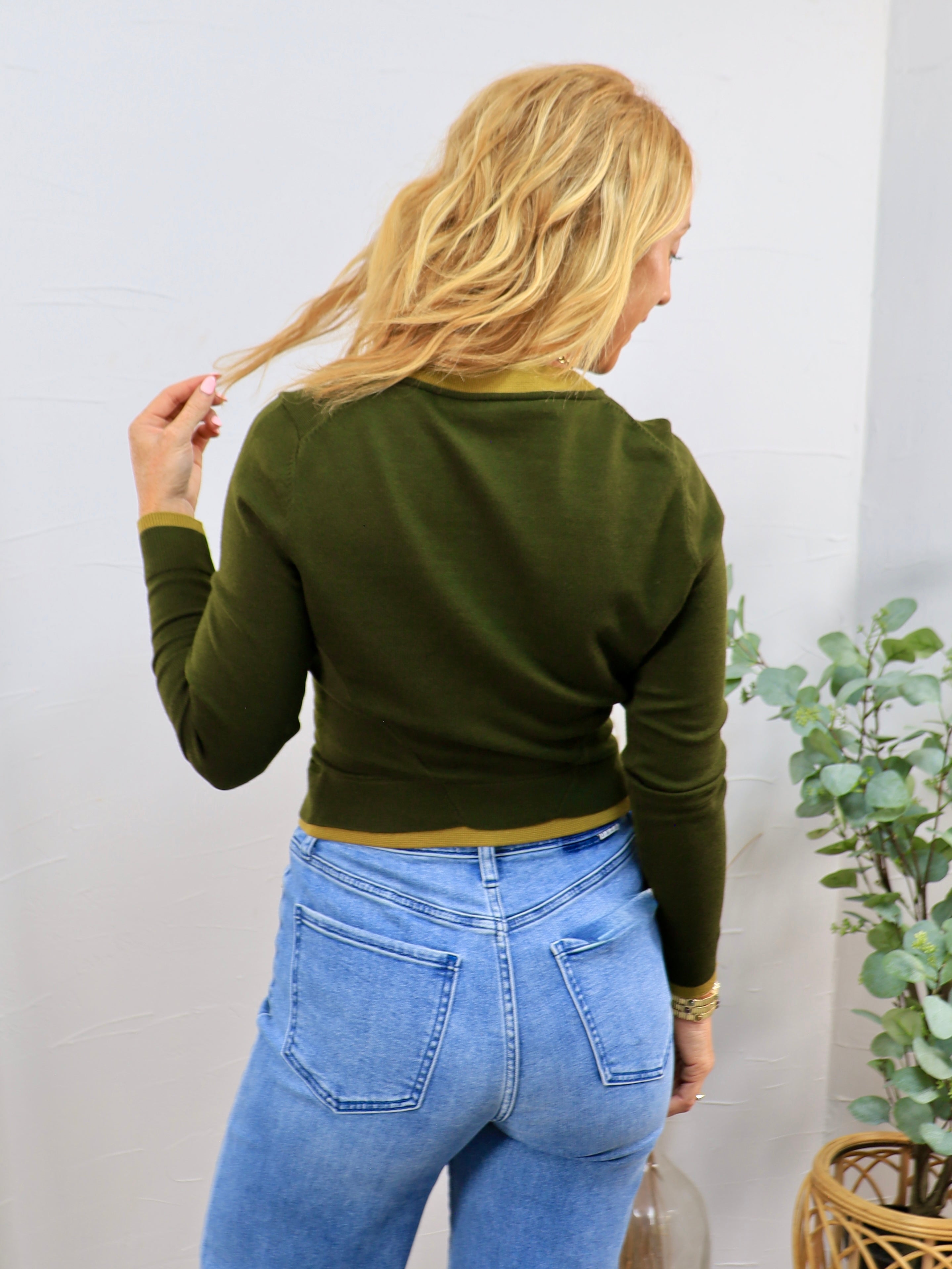 Olive green cropped cardigan with mustard trim detailing, worn over light wash blue jeans, back view.