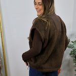 Brown shearling fleece vest with smooth sleeves, worn over dark jeans, side drawstring detail visible.