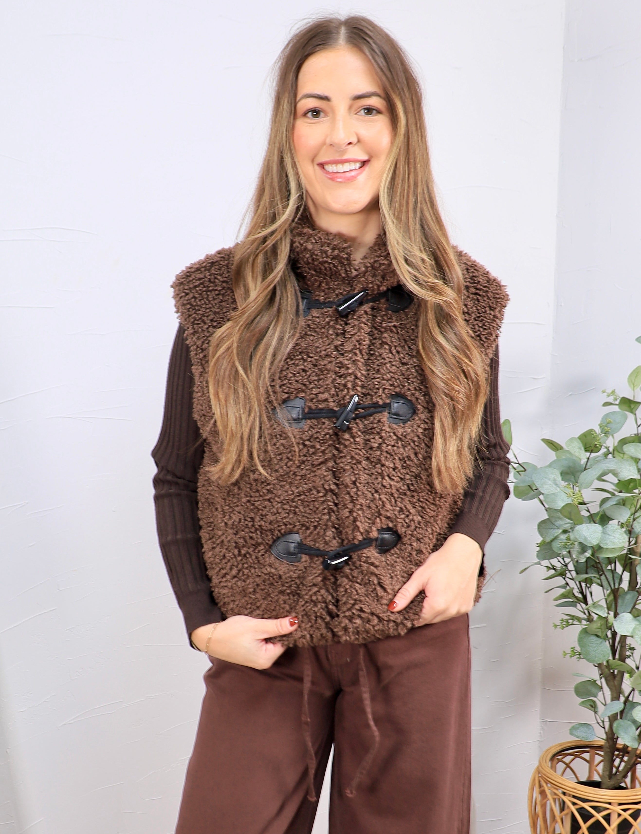 Woman wearing a chocolate brown sherpa teddy vest with black toggle closures over a ribbed dark brown top