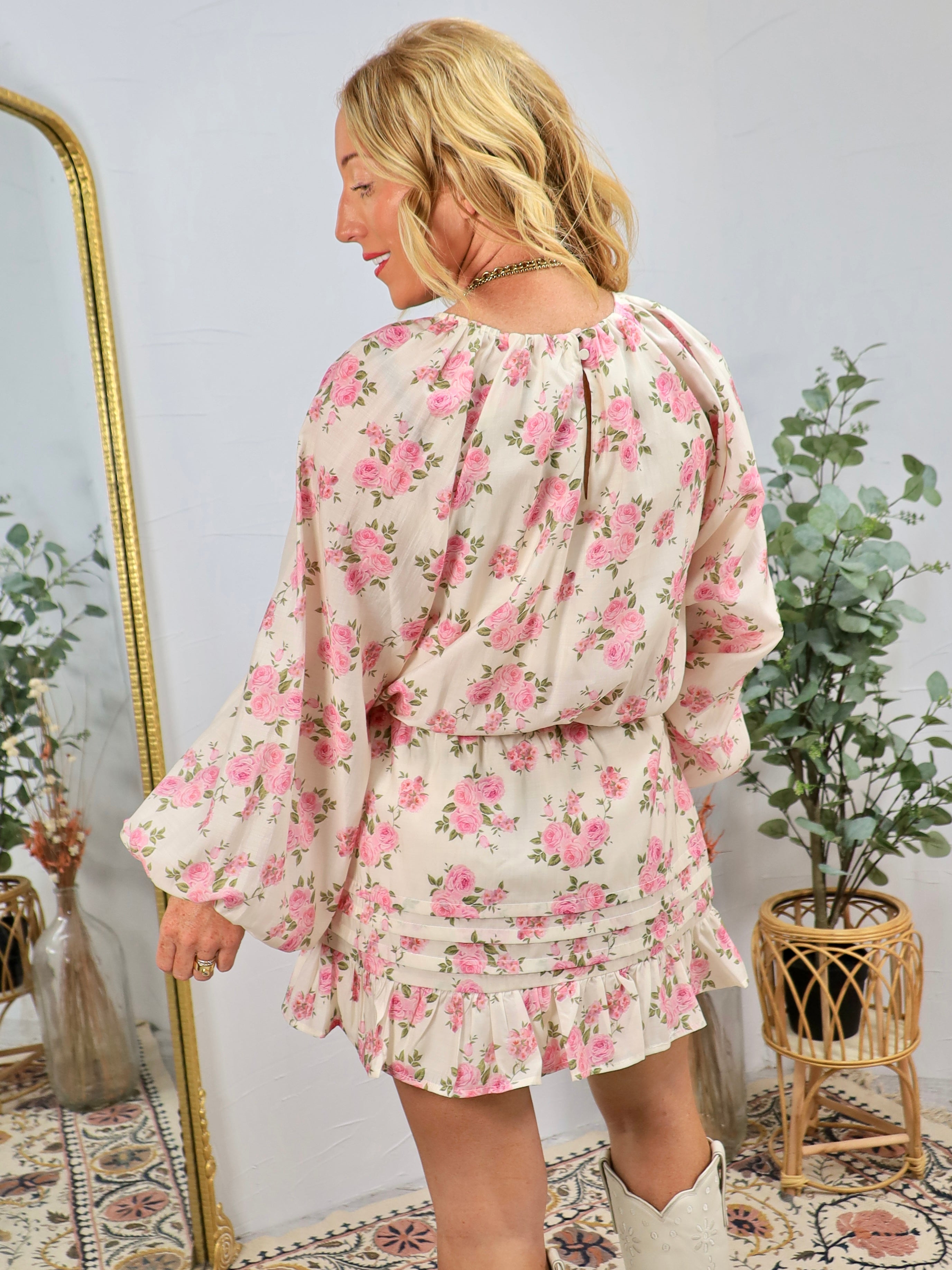 Woman wearing cream floral blouse with pink roses, long sleeves, and ruffled hem, styled with white boots in boho interior se